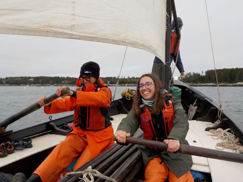 The image shows two people rowing a boat. They are both wearing orange outfits and life jackets. The person in the front is smiling. There is another person in the background. The boat has a sail. The water is calm and the sky is overcast.